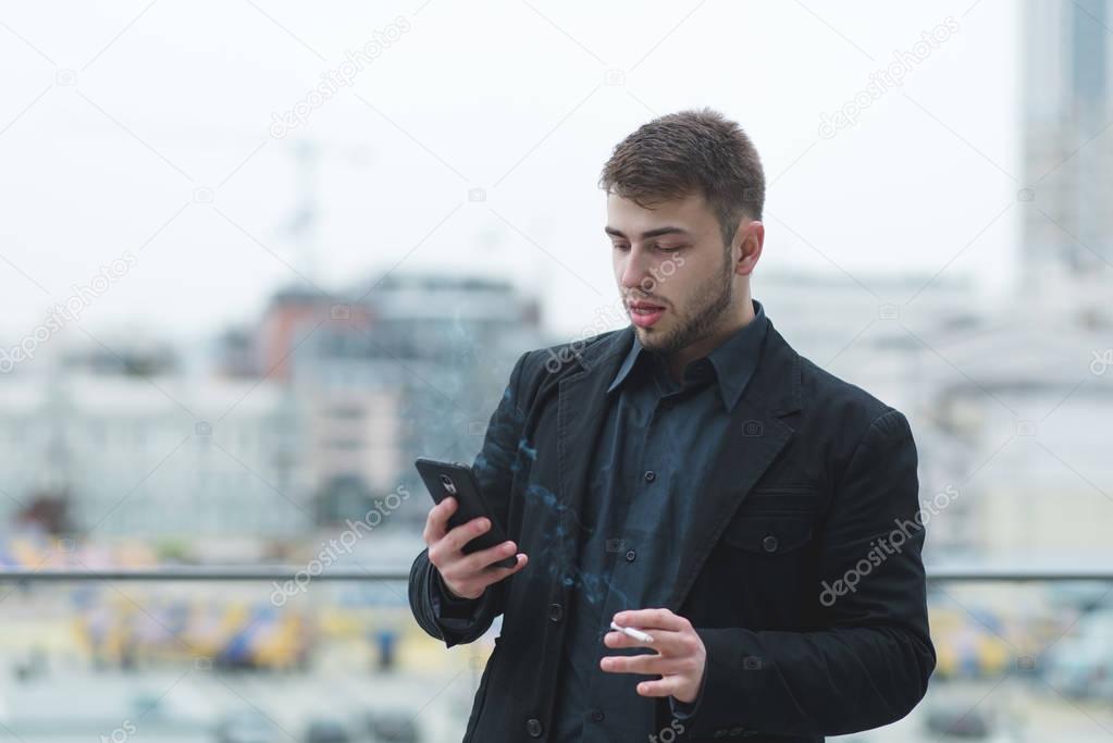 Street portrait of a man with a beard who smokes a cigarette and uses a smartphone on the street background.