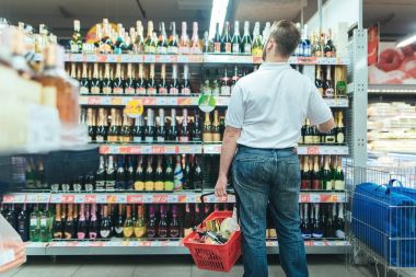 Portrait of a man standing on the background of alcoholic beverages of the supermarket. A man with a red shopping basket in his hand selects the products in the supermarket clipart