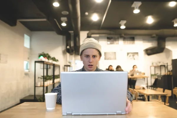 Un joven está trabajando en un café y parece sorprendido por la pantalla del portátil. El estudiante utiliza Internet en un acogedor café . —  Fotos de Stock