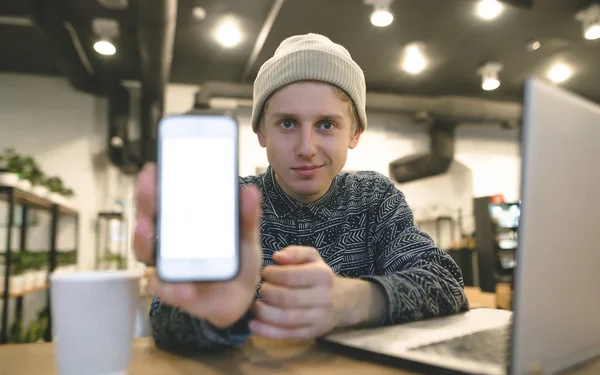 Un joven hipster muestra una pantalla de teléfono a la cámara. Una mirada a la cámara. Un estudiante trabaja para un portátil en un acogedor café . — Foto de Stock