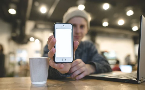 Ein junger Mann zeigt der Kamera einen Handybildschirm. konzentrieren Sie sich auf Ihr Handy. ein Telefon mit weißem Bildschirm in den Händen eines jungen Mannes. Arbeit in einem Café. — Stockfoto