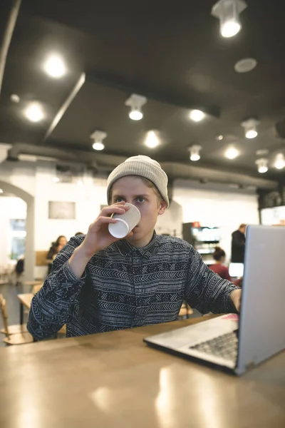 Hipster bebe café mientras trabaja en una computadora en un acogedor café. Freelancer trabaja para el café. El estudiante usa internet en la cafetería —  Fotos de Stock