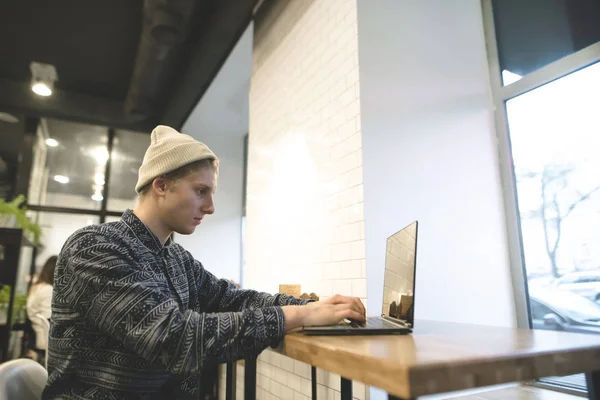 Un joven mira el monitor de su ordenador y trabaja. Estudiante mantiene la computadora en un acogedor café . —  Fotos de Stock