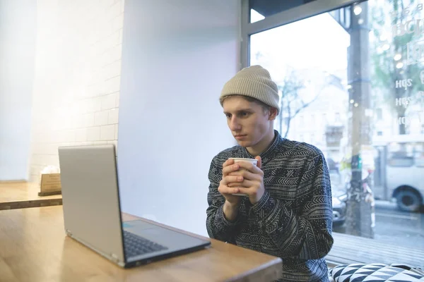 Um jovem está sentado em um café acolhedor com uma xícara de café em suas mãos e assistindo a um vídeo em um laptop. Estudante elegante em um computador em um café . — Fotografia de Stock