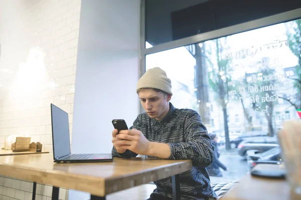 Sitzt ein stylischer junger Mann in einem gemütlichen Café neben einem Laptop und benutzt ein Smartphone. Ein junger Mann arbeitet in einem Café. — Stockfoto