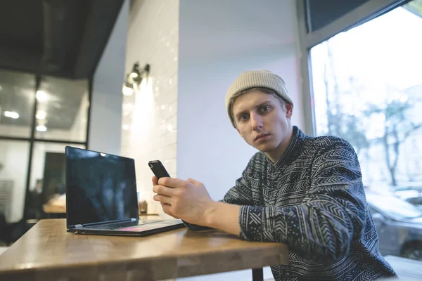 Hipster se sienta en un acogedor café cerca de un ordenador portátil, utiliza un teléfono inteligente y mira a la cámara. Un joven serio en un acogedor café con una cafetería cerca de la computadora portátil . —  Fotos de Stock