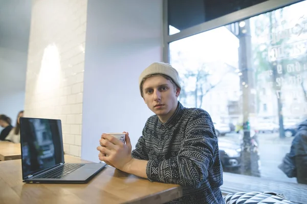 Un joven con estilo con un vaso de café en las manos se sienta en un acogedor café cerca de la computadora portátil. Una mirada a la cámara . — Foto de Stock