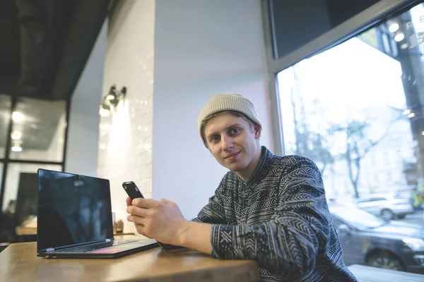 Un estudiante positivo sentado en un acogedor café con un portátil y un teléfono en sus manos. Hipsters mira a la cámara y sonríe . —  Fotos de Stock