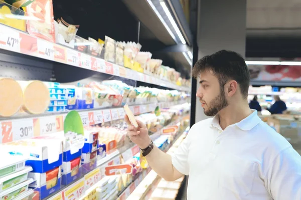 Un hombre con barba compra comida en un supermercado. Un joven selecciona productos lácteos en los estantes del supermercado . — Foto de Stock