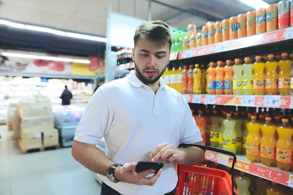 A man with a basket looks at the phone while shopping at a supermarket. Shopping in the store. is in the supermarket near the drinks. — Stock Photo, Image