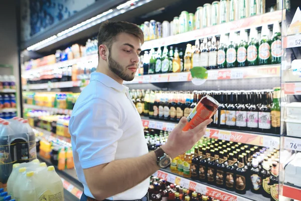 A handsome bearded man chooses beer in a supermarket. The buyer buys alcohol at a supermarket. A man looks at a can of beer — Stock Photo, Image