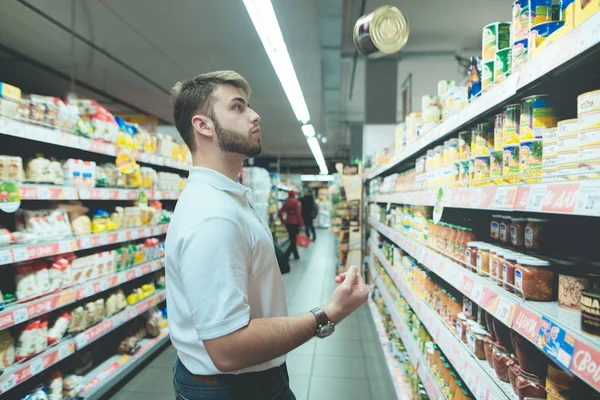 Un bărbat frumos alege conservele din rafturile supermarketurilor. Un om cu barbă jonglează cu bunurile din magazin. . — Fotografie, imagine de stoc