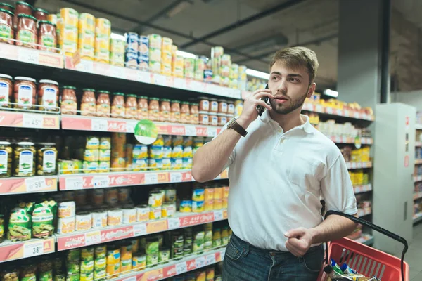 Sakallı yakışıklı bir adam bir süpermarkette telefonla konuşuyor. Kırmızı-sepet alıcı vasıl belgili tanımlık stok ürünler seçer ve telefon üzerinden iletişim kurar. — Stok fotoğraf