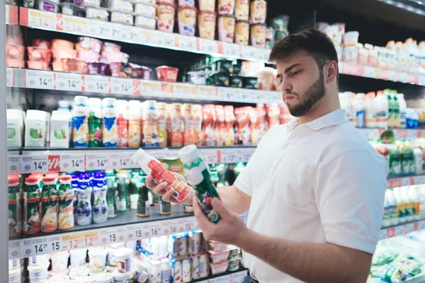 Um homem emocional escolhe iogurte no departamento de leite do supermercado. O homem comprador compra os produtos na loja. Seleção de produtos no supermercado . — Fotografia de Stock