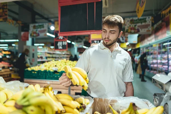 Un guapo barbudo compra plátanos en un supermercado. Un hombre elige fruta en el departamento de verduras del supermercado. Compras en la tienda . — Foto de Stock