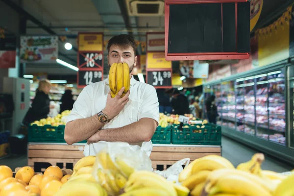 Retrato de un hombre con plátanos alrededor de su cara mientras compraba en un supermercado. Un hombre con plátanos en sus manos mira a la cámara en el fondo del departamento de verduras del supermercado — Foto de Stock