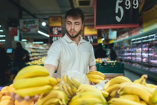 A man with a beard chooses bananas in the vegetable department of the supermarket. A man buys fruit in a store. Purchase of products in a supermarket. — Stock Photo, Image