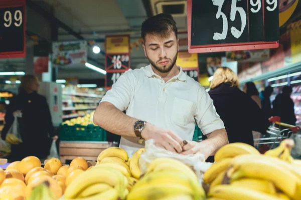 Un hombre guapo compra fruta en un supermercado. Un hombre elige plátanos en la tienda. Compra de productos en un supermercado . — Foto de Stock