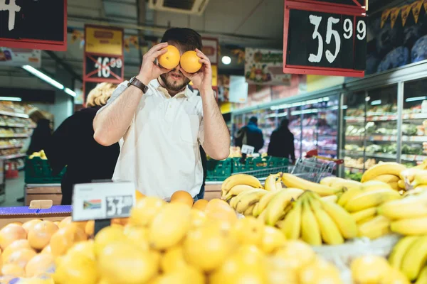 Retrato de un hombre alegre en el departamento de verduras del supermercado. Un hombre gracioso sostiene naranjas alrededor de los ojos. Compra de fruta en un supermercado . — Foto de Stock