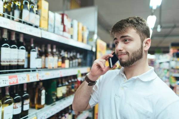 Uśmiechnięty mężczyzna wybiera winel w supermarkecie i rozmowy przez telefon. Wybór towaru w supermarkecie. Zakupy w sklepie — Zdjęcie stockowe