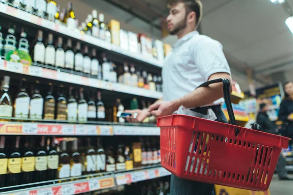 Een man met een mandje product kijkt naar het schap van de supermarkt en wijn kiest. Een man maakt aankopen in de winkel. Focus op de mand. — Stockfoto