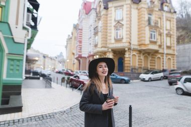 Portrait of a stylish smiling woman walking around a beautiful old town with a cup of hot drink in her hands. Girl hipster on the background of a beautiful city street. clipart