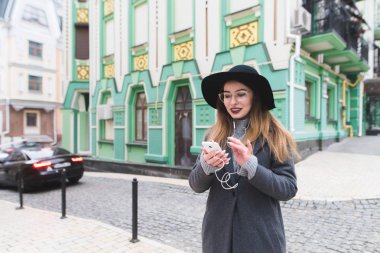 Şık bir kadın telefon eski şehrin güzel mimari arka plan üzerinde kullanır. Kız şehir etrafında yürür ve kulaklık sokakta müzik dinler.