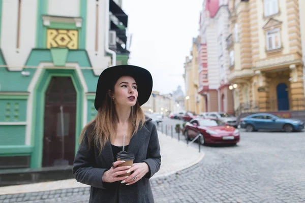 Portræt af en stilfuld smuk kvinde med en kop kaffe i hånden mod en smuk by. En smuk kvinde går rundt i byen med et glas kaffe - Stock-foto