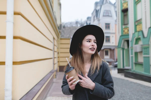 Portrait of a positive girl with a cup of drinks in her hands against a background of beautiful colored architecture. Happy girl walking along the streets of the old town. — Stock Photo, Image