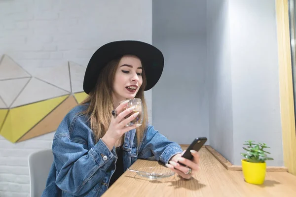 Porträt eines stilvollen und positiven Mädchens mit einem Smarthon in der Hand, das in einem gemütlichen Café ein Heißgetränk trinkt. Kaffeepause. Blick auf das Smartphone während des Mittagessens. — Stockfoto