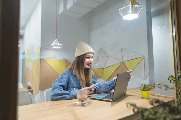 Hipster menina senta-se em um café acolhedor com um laptop e sorri. Retrato de uma menina bonita através da janela do café . — Fotografia de Stock