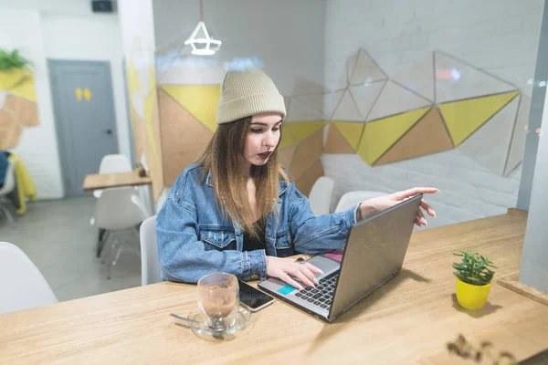 Una bella ragazza freelance lavora su un computer portatile in un elegante caffè di design. Una donna elegante utilizza un computer in un caffè per una tazza di caffè . — Foto Stock