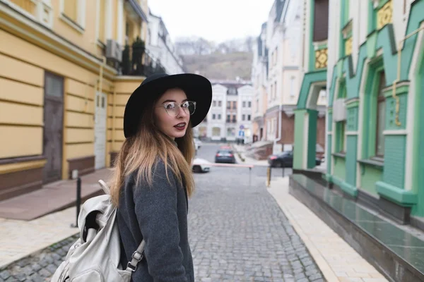 Une fille élégante en manteau et chapeau se promène dans la vieille ville et regarde la caméra. Une belle femme voyage dans une belle ville — Photo