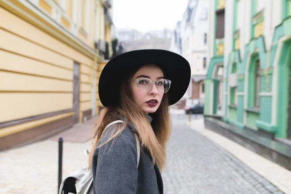 Portrét krásné stylové ženy v čepici a brýle na pozadí ulice starého města. Pohled na kameru. — Stock fotografie