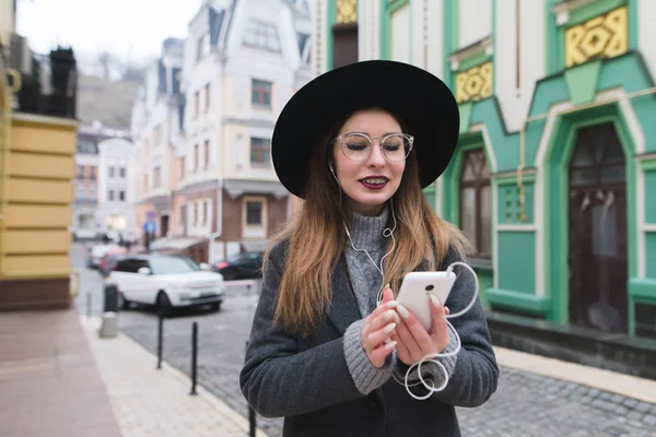Позитивна, стильна жінка слухає музику, дивиться в телефон і посміхається на тлі вулиці старої спарчки. Прогулянка в прекрасному місті . — стокове фото