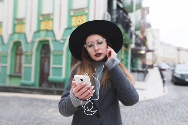 Μια κομψή γυναίκα ακούει μουσική στα ακουστικά για το ιστορικό της μια όμορφη παλιά πόλη και διακόπτες τραγούδια στο τηλέφωνο. Εστίαση στο τηλέφωνό σας. — Φωτογραφία Αρχείου