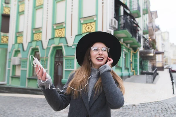 A cheerful woman with a phone in her hands and headphones in her ears smiles against the backdrop of beautiful architecture. Street portrait of a happy girl. — Stock Photo, Image