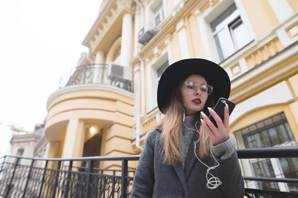 Ein Hipster-Mädchen mit Hut und Brille blickt auf das Telefon vor dem Hintergrund eines schönen Hauses. Straßenporträt einer stilvollen Frau vor dem Hintergrund der Architektur der Altstadt — Stockfoto