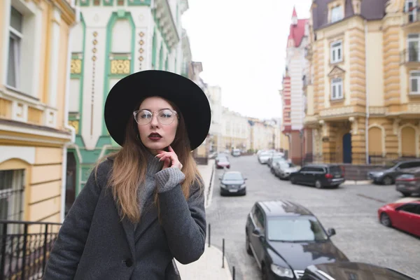 Stylische junge Frau posiert vor der Kulisse einer schönen alten Stadt. im herbstlich gekleideten Mädchen-Hipster steht auf dem Hintergrund der alten Straße. — Stockfoto