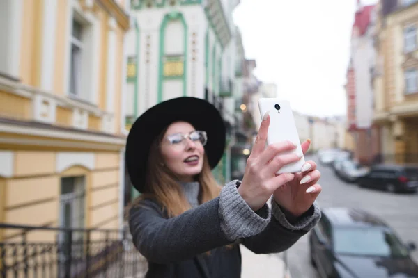 Fata Hipster face selfie pe telefonul tau in timp ce merge in frumosul oras vechi. Celph pe fundalul unei frumoase străzi vechi. Concentrează-te pe telefon . — Fotografie, imagine de stoc