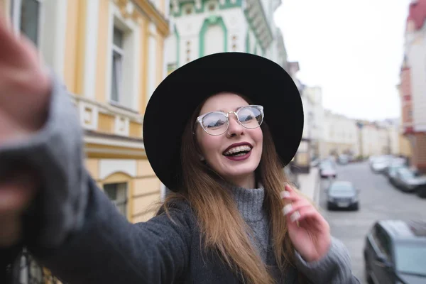 Stylish girl with braces takes selfie krasvoho against the background of the urban landscape. The girl tourist on background makes selfie beautiful streets of the old town. — Stock Photo, Image