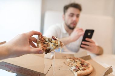 Bir parça pizza onun elinde. Gençler kutusundan pizza yemek. Koca bir cep telefonu kullanan ve pizza yiyor.