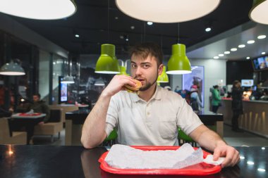 Bir adam hızlı bir kundak restoranda bir masada oturuyor, lezzetli yemek yeme ve kameraya bakıyor. Fast food kavramı.