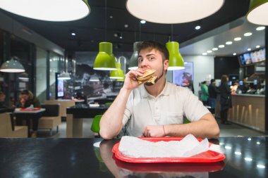 Komik bir adam bir restoranda lezzetli bir hamburger yiyor ve kameraya benziyor. Fast Food yemek.