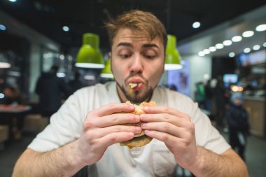 Bir sakallı aç adam iştahla hamburger yiyor. Bir sandviçi arıyorsunuz komik bir adam. Burger üzerinde odaklanın. Fast food kavramı.