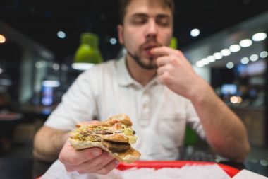 Genç bir adam elinde büyük iştah açıcı sandviç. Adam fast food Restoran beslenir. Yetenekli burger yakın çekim.