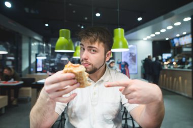 Yakışıklı bir adam elinde burger Puan onun parmak ve kameraya benziyor. Adam fast food restoranda yiyor..