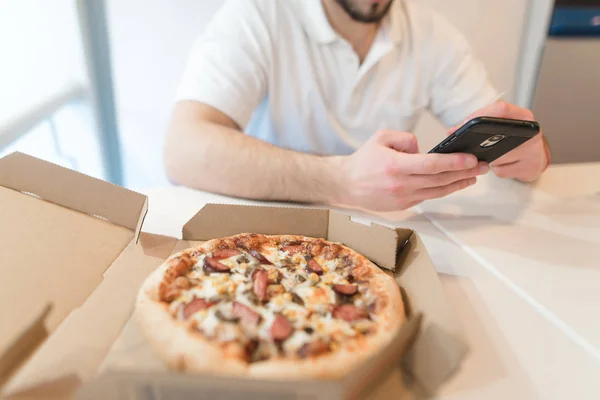 Uma caixa de pizza apetitosa está sobre a mesa. Um homem usa um telefone no fundo de uma caixa aberta de pizza saborosa. Foco na pizza — Fotografia de Stock