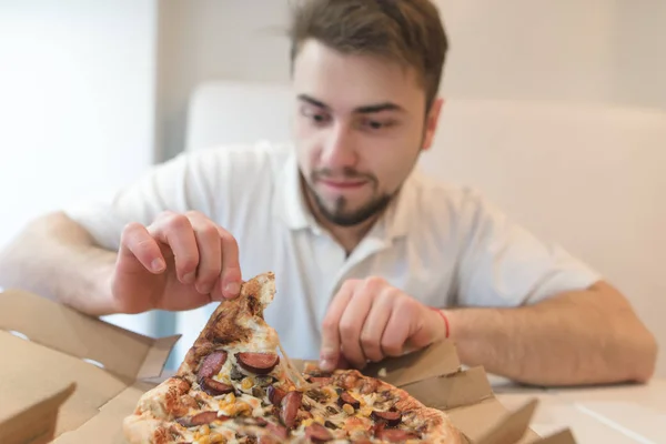 Um homem bonito com um olhar faminto aperta um pedaço de pizza fora da caixa e está prestes a comê-lo. Almoço com uma pizza quente apetitosa no restaurante . — Fotografia de Stock