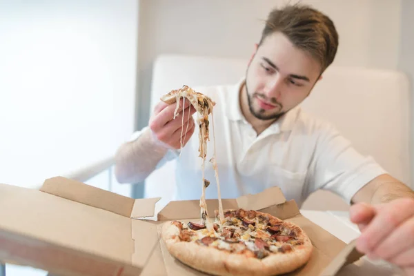 Um homem bonito segura uma fina pizza apetitosa em suas mãos e olha para ela com um olhar faminto. O homem come uma pizza quente de uma caixa de papelão . — Fotografia de Stock